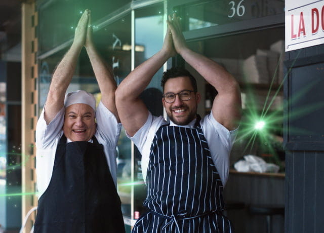 Two men standing in front of their restaurant happily with their hands up