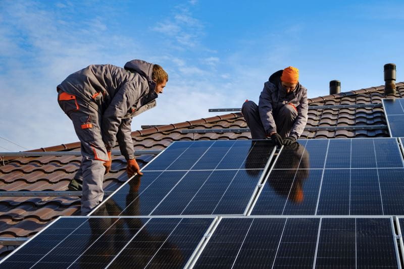 Two men are fitting solar panels onto the roof of a house